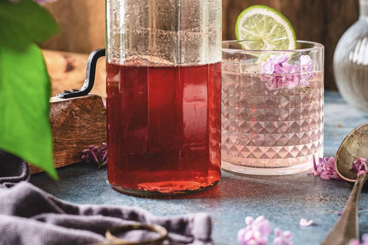 A glass pitcher of red liquid beside a glass with pink drink made with lilac syrup, lilac flowers, and a lime slice garnish.