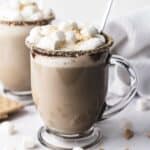 A glass mug of hot chocolate topped with whipped cream and marshmallows, with a graham cracker rim&mdash;just like a cozy S&rsquo;mores Latte Recipe.