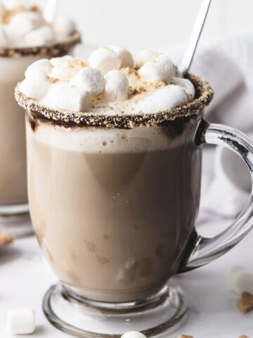 A glass mug of hot chocolate topped with whipped cream and marshmallows, with a graham cracker rim&mdash;just like a cozy S&rsquo;mores Latte Recipe.