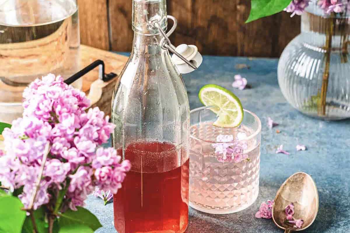 A glass bottle of pink drink made with lilac syrup and a glass with lime, surrounded by pink flowers on a blue tabletop.