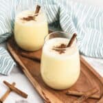 Two glasses of creamy eggnog topped with cinnamon sticks and sprinkled cinnamon sit on a wooden tray, with a striped cloth nearby.