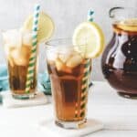Two glasses of homemade sweet tea with lemon slices and striped straws, with a pitcher of iced tea in the background.