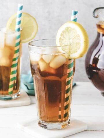 Two glasses of homemade sweet tea with lemon slices and striped straws, with a pitcher of iced tea in the background.