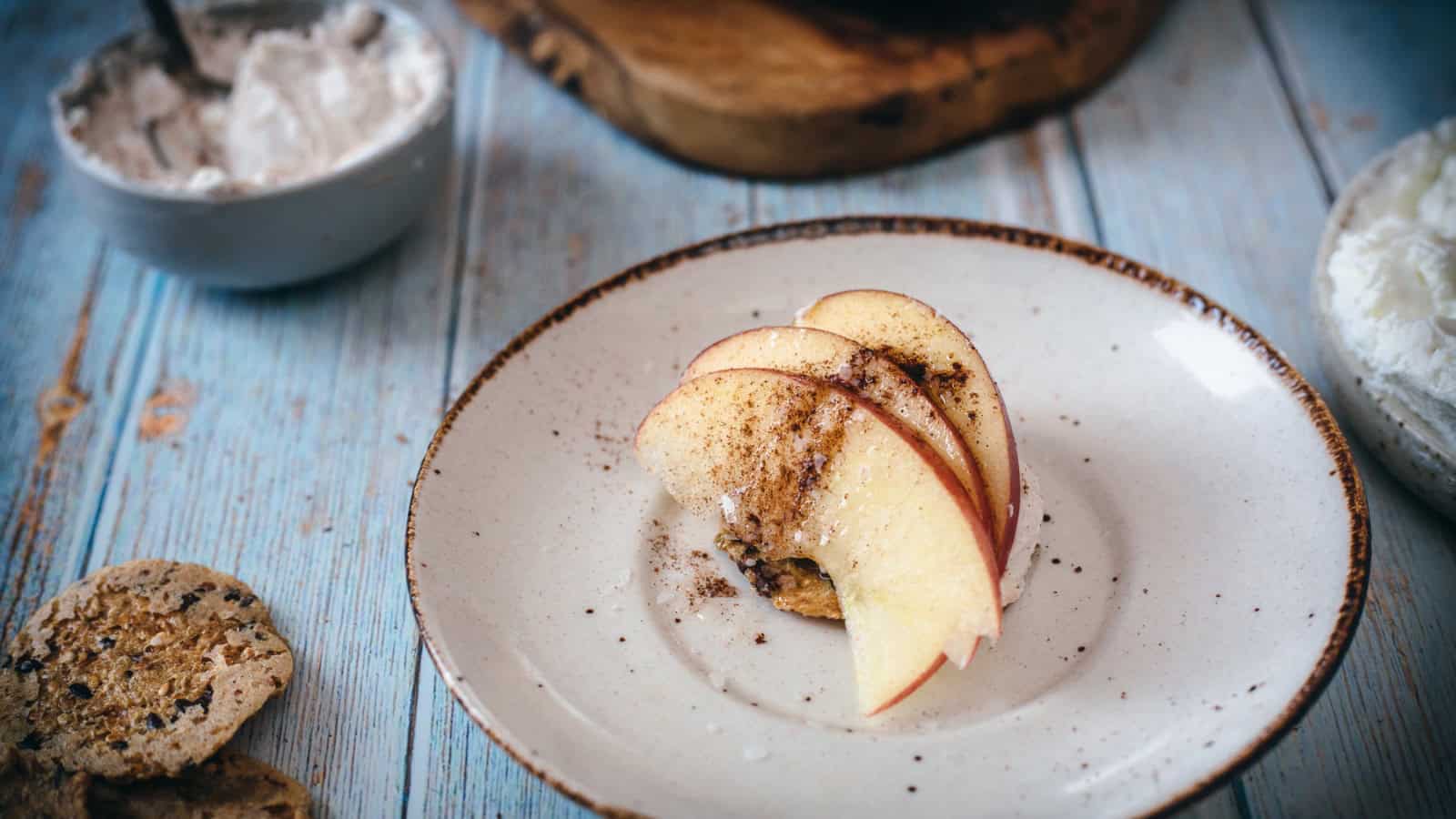 A white plate with thin apple slices sprinkled with cinnamon. A crumbled cookie is partially visible on the left side of the light blue wooden table.