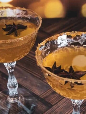 Two ornate glasses filled with an amber cocktail, garnished with star anise and a sugared rim on a wooden table.