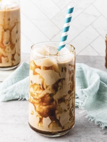 A glass of iced caramel coffee with caramel drizzle and a striped straw sits on a light surface with a blue cloth.