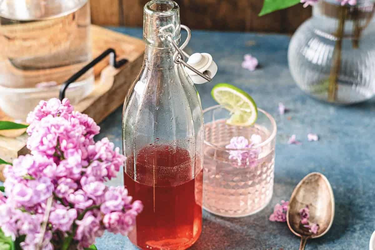 A bottle of pink lilac syrup beside a glass with lime slice, lilac flowers, and a spoon on a blue surface.