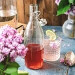 A glass bottle of pink lilac syrup drink and a glass with a lime slice, surrounded by lilac flowers on a blue surface.
