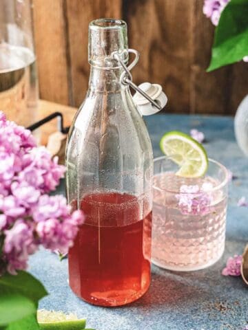 A glass bottle of pink lilac syrup drink and a glass with a lime slice, surrounded by lilac flowers on a blue surface.
