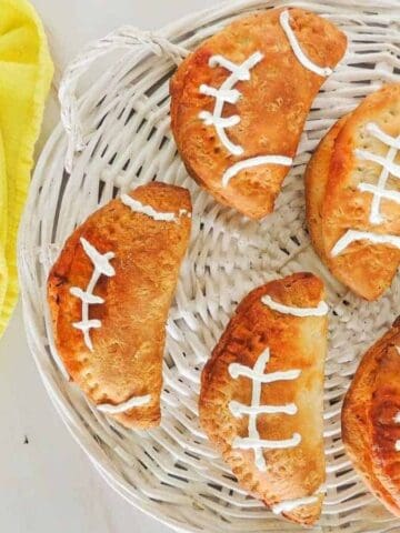 Football-shaped empanadas with white icing laces on a wicker tray and yellow napkin.