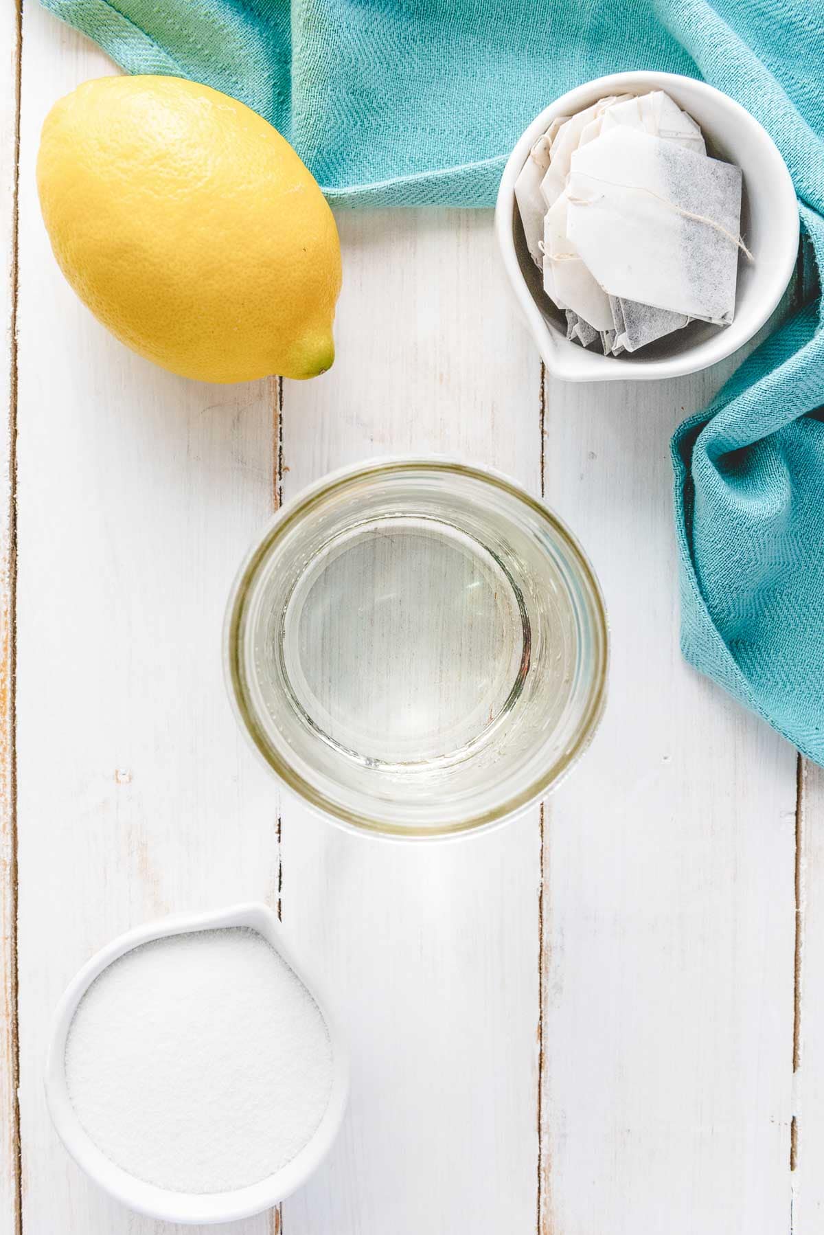Flat lay of a lemon, bowl of tea bags, sugar, and a glass of water on a white wooden surface with a teal cloth&mdash;perfect essentials for crafting homemade sweet tea.