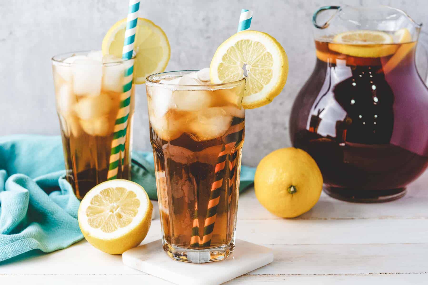Two glasses of homemade sweet tea with lemon slices and straws, a pitcher of iced tea, and fresh lemons sit on a table.