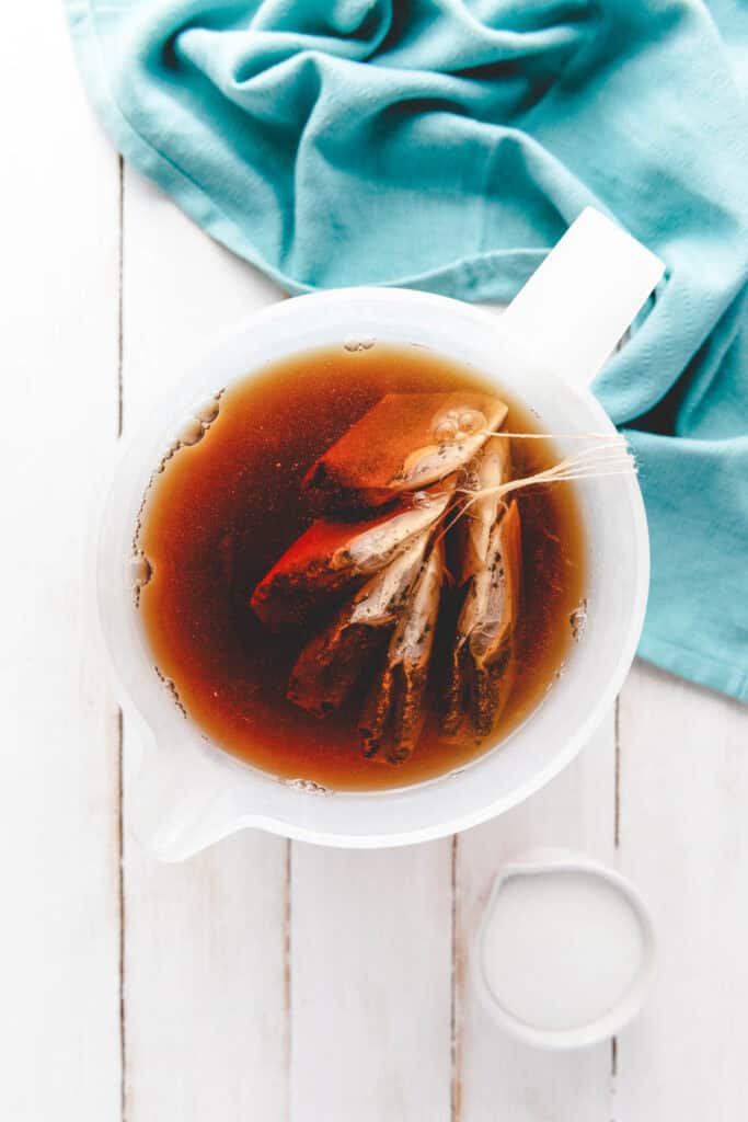 A white pitcher with homemade sweet tea and tea bags steeping in dark tea, next to a small bowl of sugar and a blue cloth.