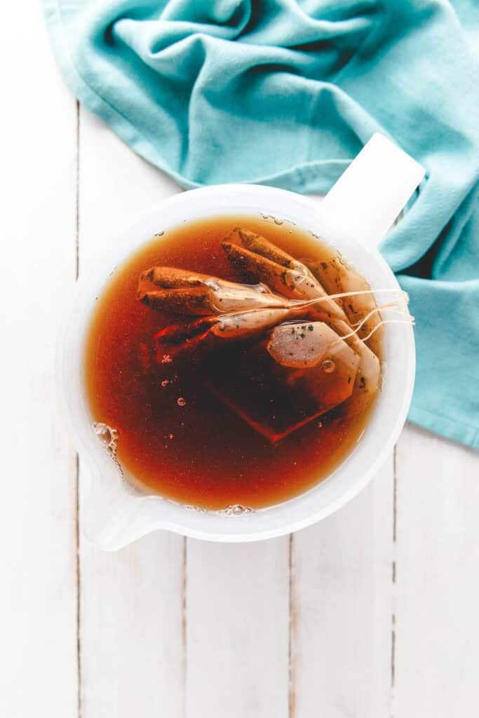 A white pitcher with homemade sweet tea and tea bags steeping in dark tea, next to a teal cloth on a white wooden surface.