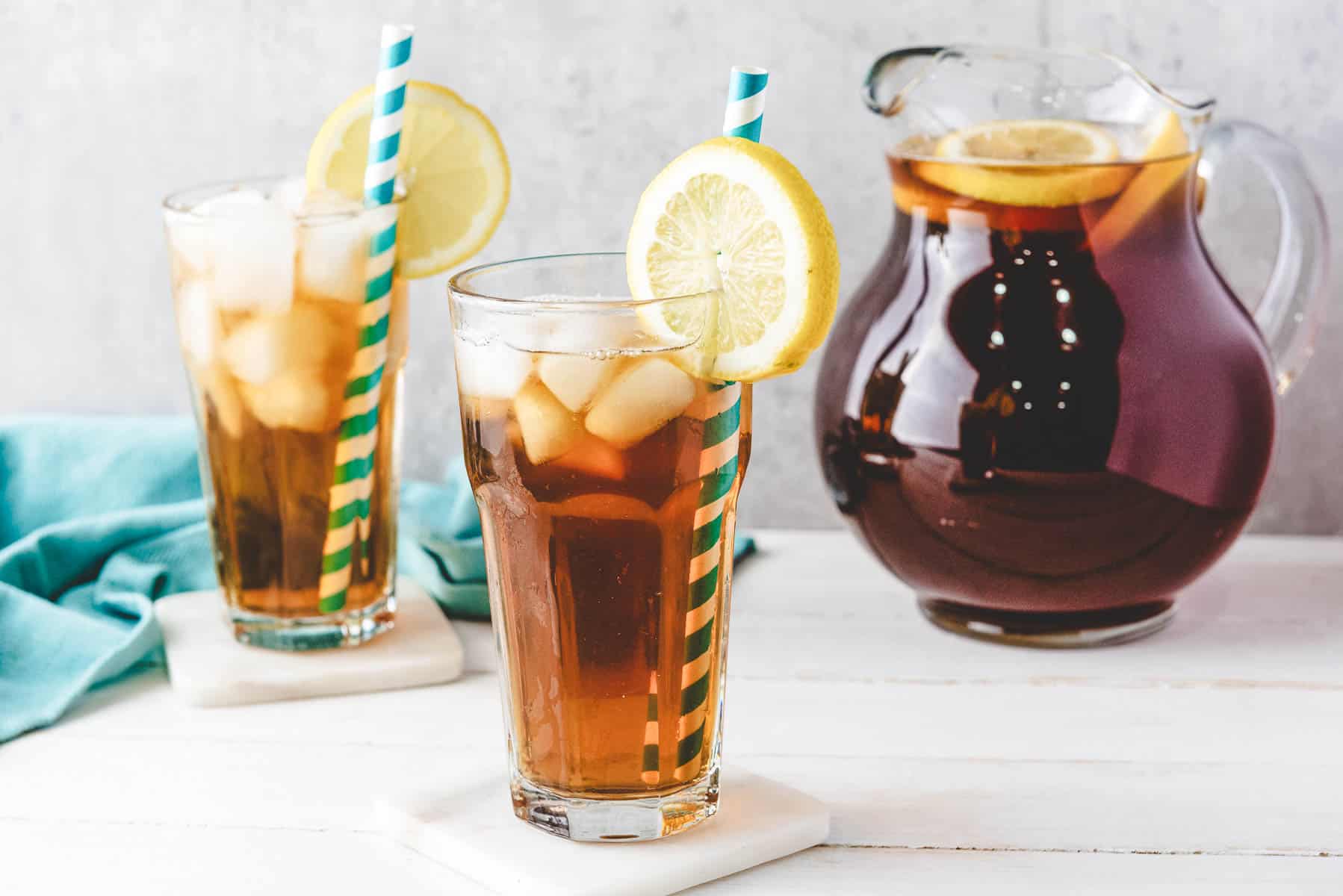 Two glasses of homemade sweet tea with lemon slices and straws sit beside a pitcher of iced tea on a white table.