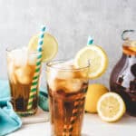 Two iced tea glasses filled with homemade sweet tea, garnished with lemon slices and striped straws, sit beside fresh lemons and a pitcher on a white table.