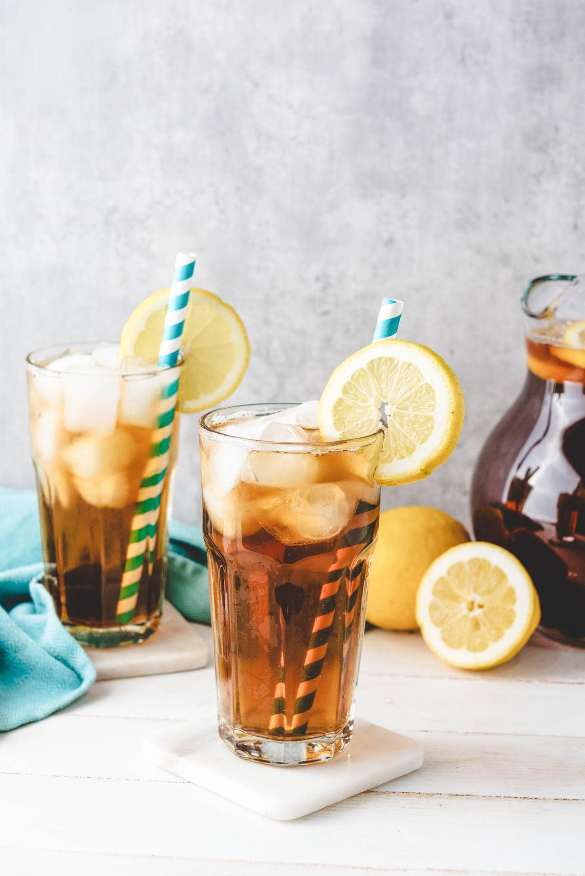 Two iced tea glasses filled with homemade sweet tea, garnished with lemon slices and striped straws, sit beside fresh lemons and a pitcher on a white table.