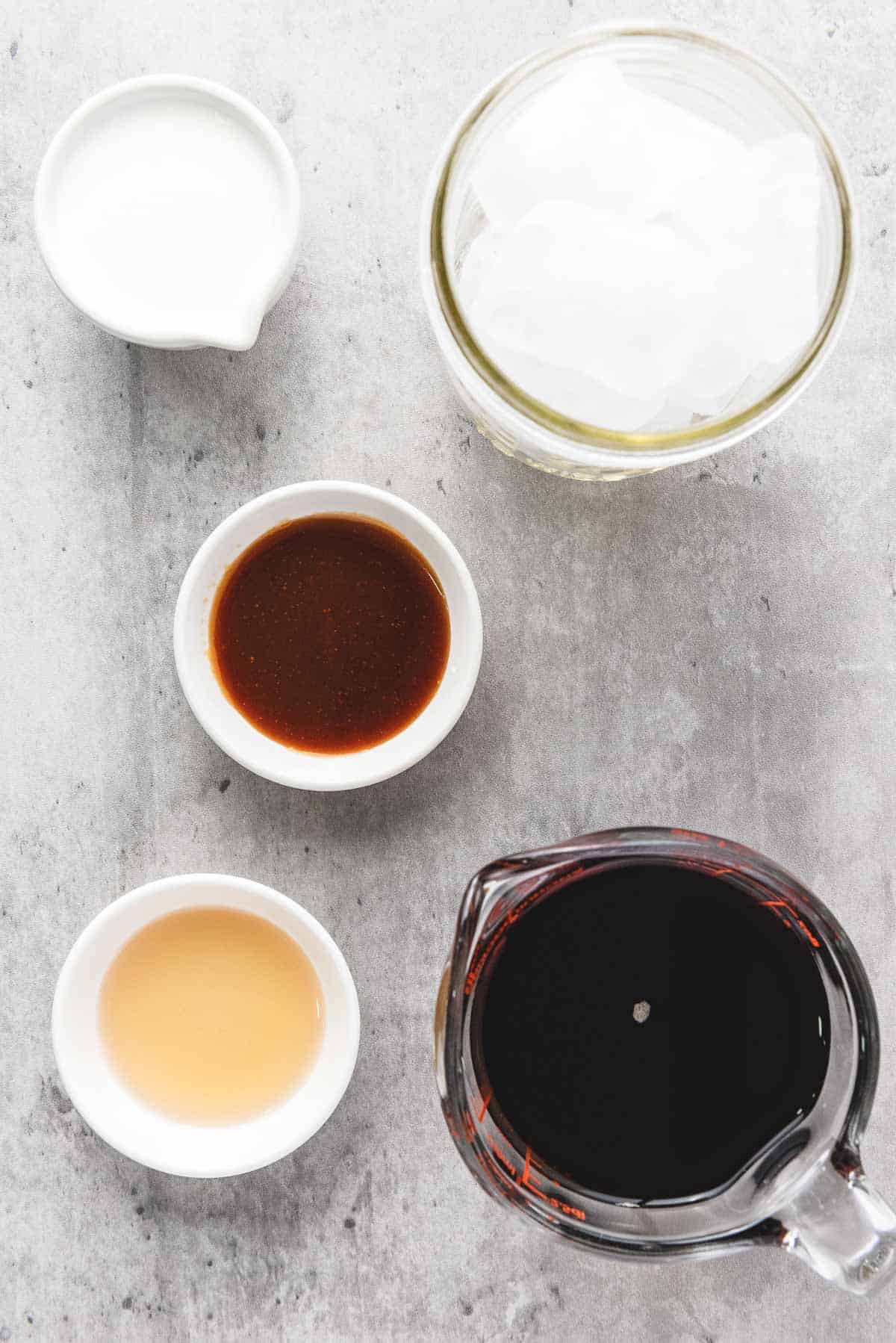 Five bowls with coconut milk, ice cubes, iced caramel coffee, light liquid, and dark sauce on a gray surface.