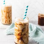 Two glasses of iced caramel coffee with caramel drizzle and blue striped straws sit on a light surface.