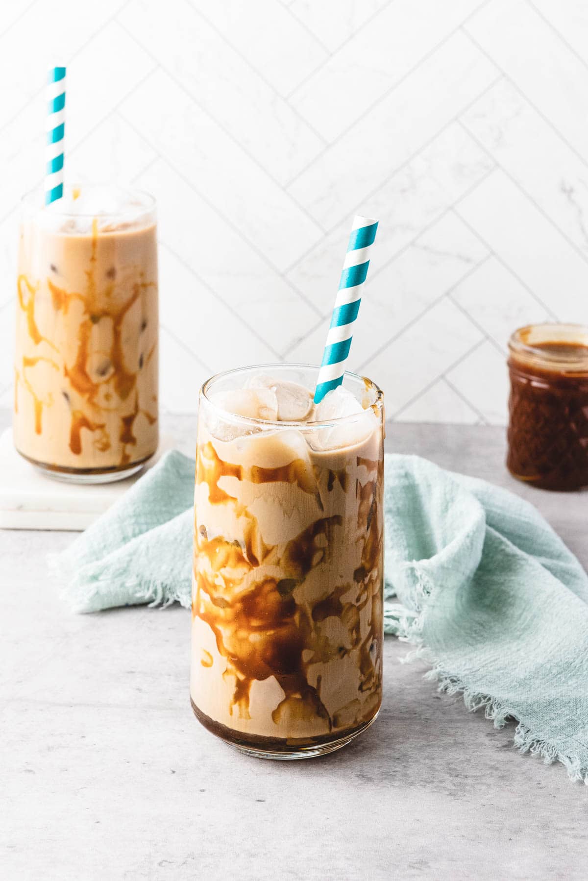 Two glasses of iced caramel coffee with caramel drizzle and blue striped straws sit on a light surface.