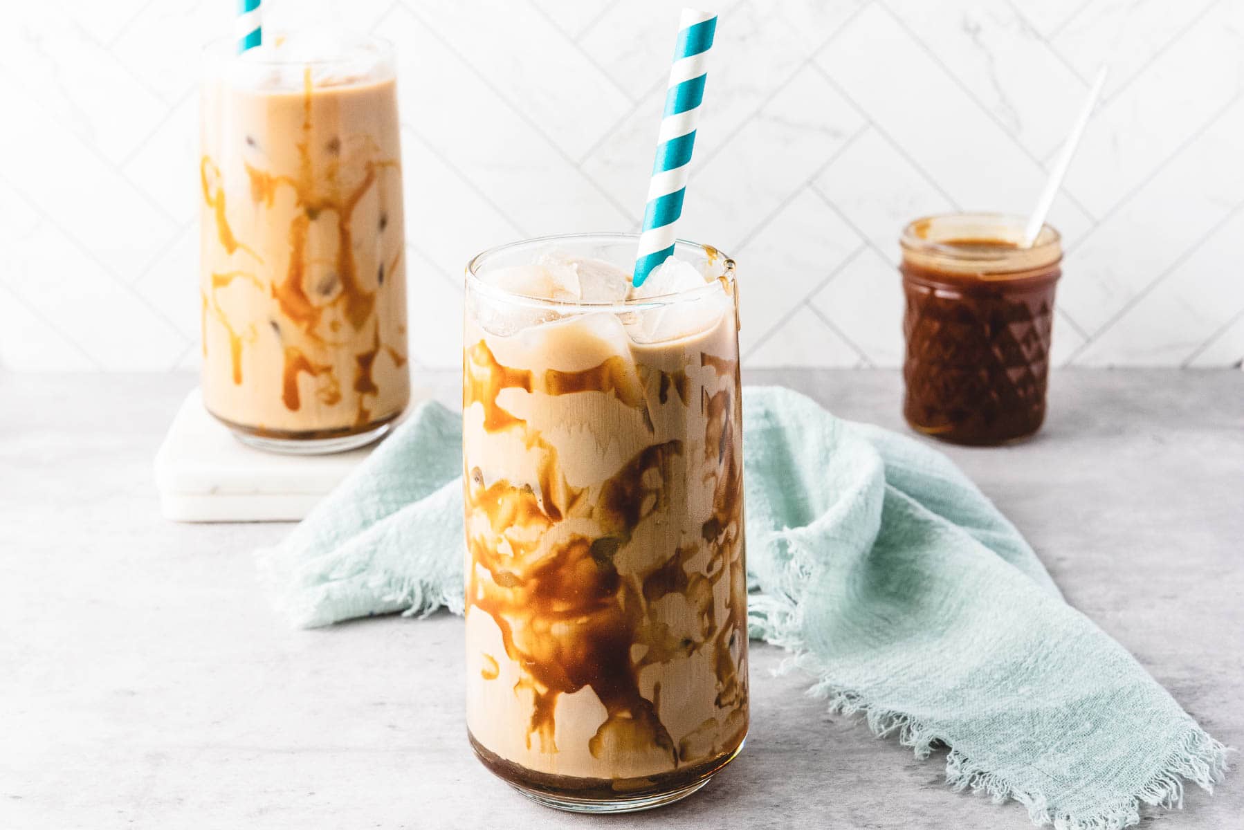 Iced caramel coffee drinks with caramel drizzle, blue striped straws, and a jar of caramel sauce are displayed on a light background.