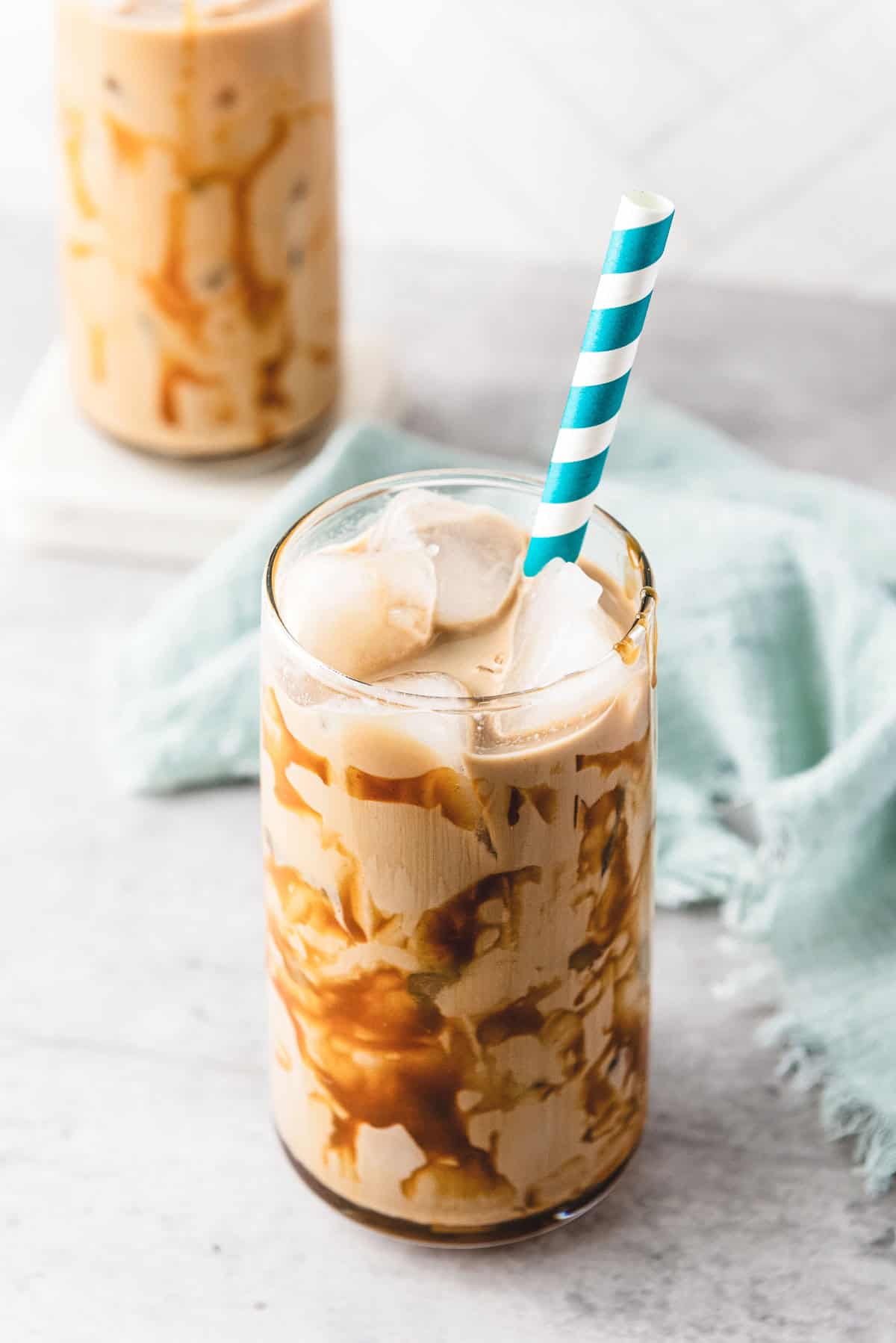 Iced caramel coffee with a caramel drizzle, ice cubes, and a blue-striped straw in a clear glass on a light surface.