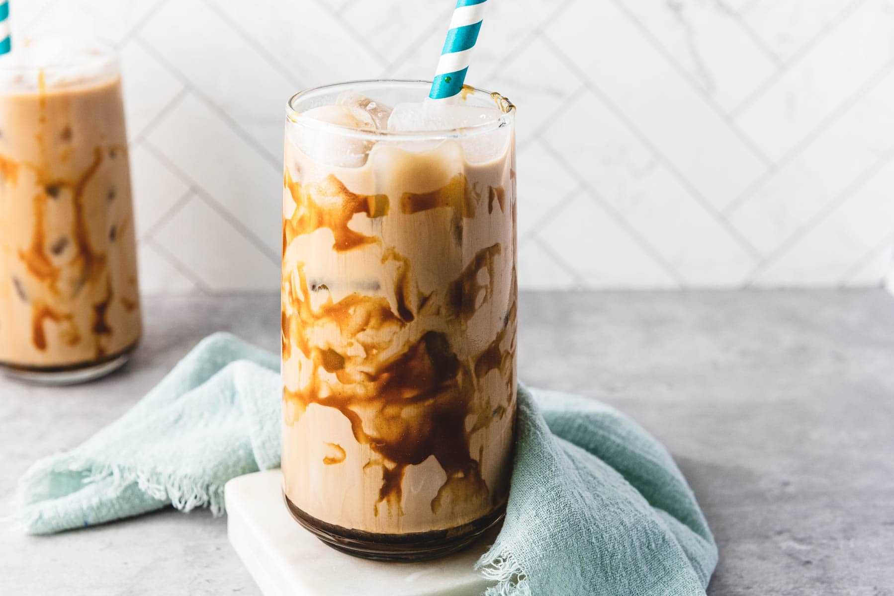 Iced caramel coffee with a caramel drizzle in a glass and a striped straw, set on a blue cloth and gray surface.