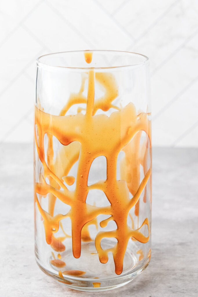 Clear glass of iced caramel coffee with caramel sauce dripped inside, standing on a gray surface with a tiled background.