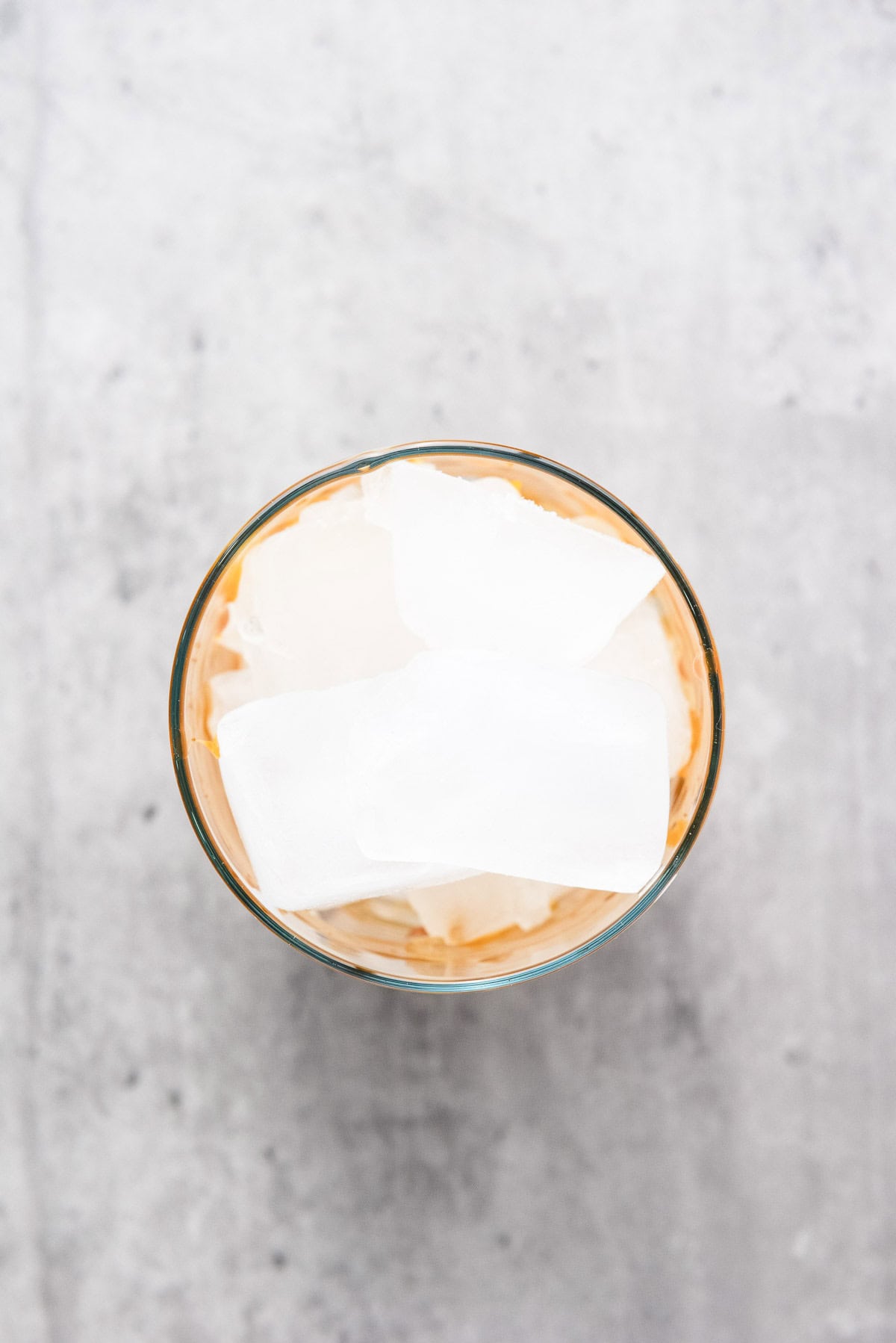 A glass of iced caramel coffee with large ice cubes sits on a light gray, textured surface, viewed from above.