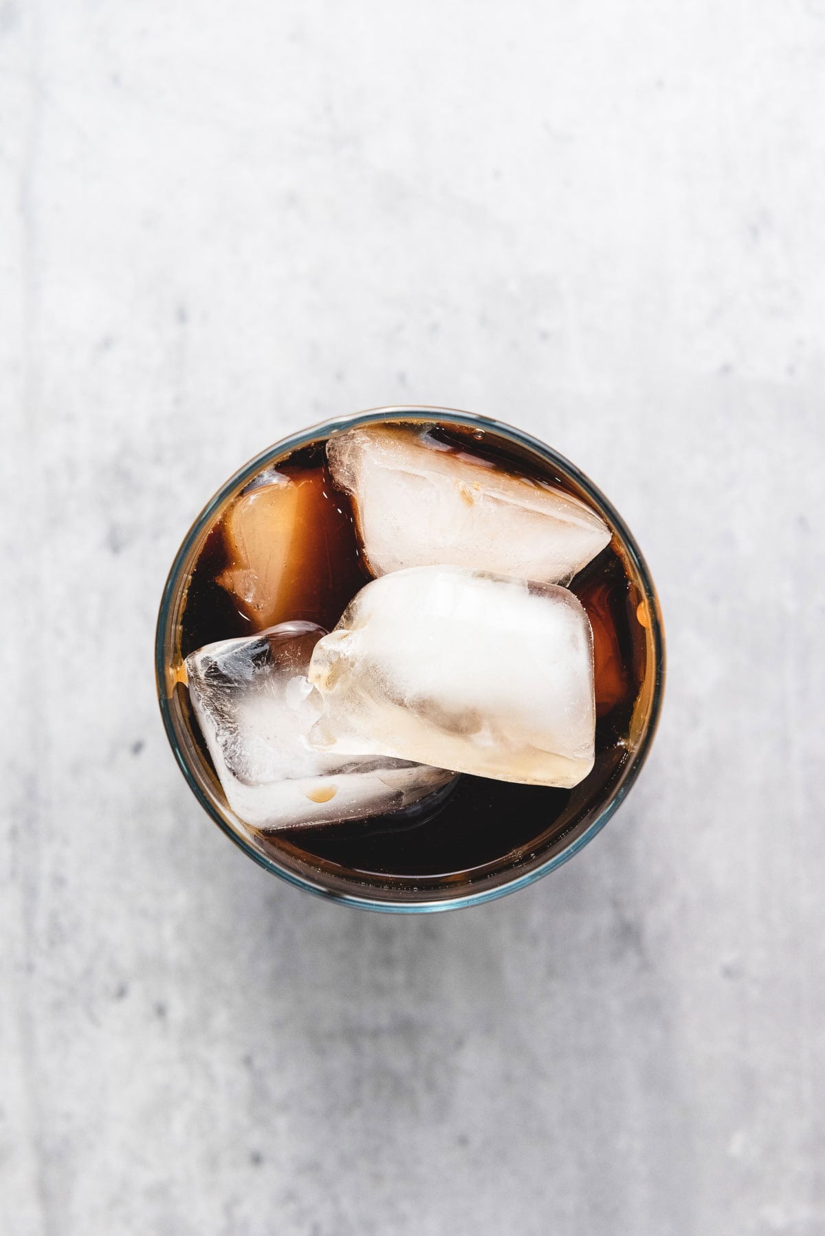 A glass of iced caramel coffee with large ice cubes is seen from above on a light gray surface.