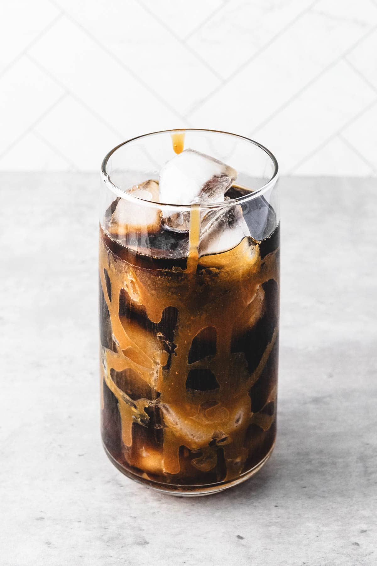 A glass of iced caramel coffee with swirls of caramel on the inside, filled with ice cubes, sits on a light surface.