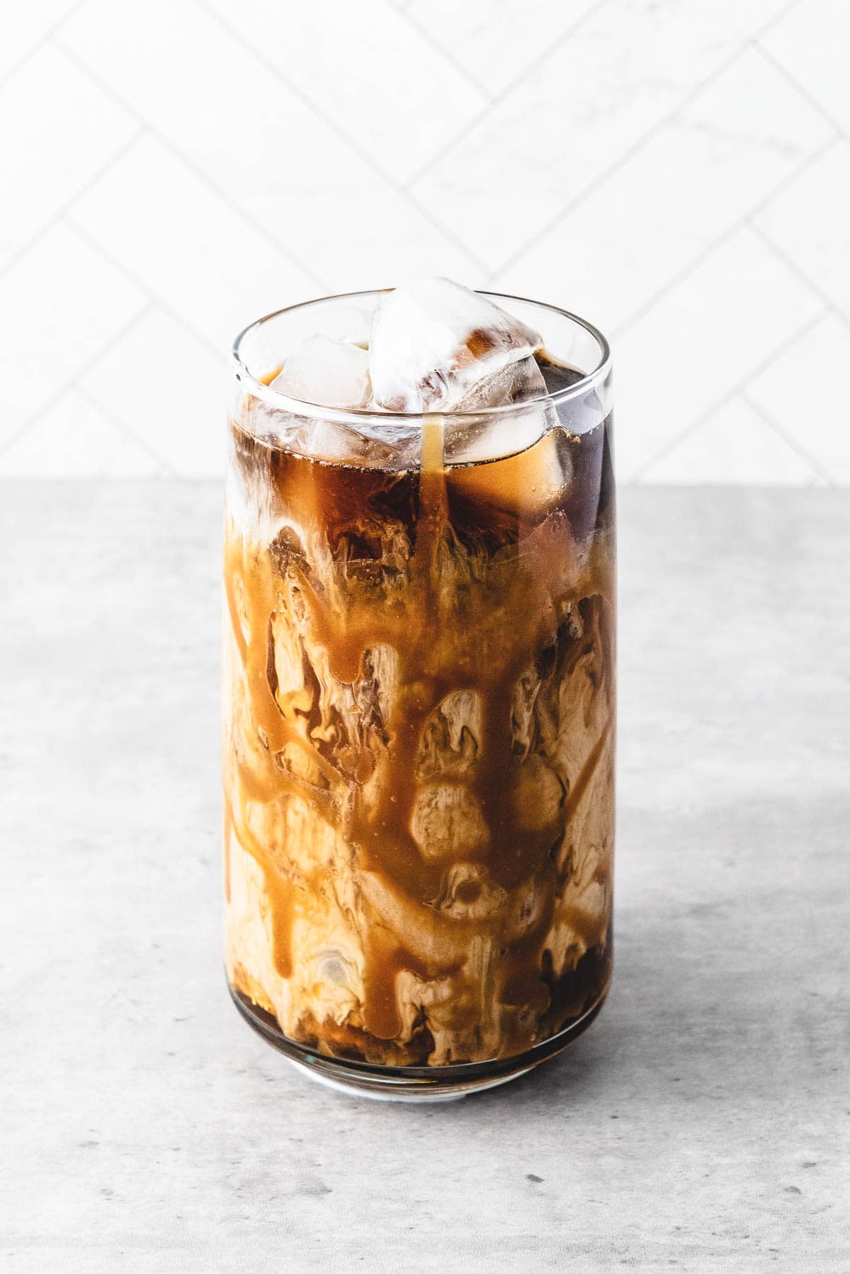 A glass of iced caramel coffee with milk swirling inside, topped with ice cubes, on a light gray surface.