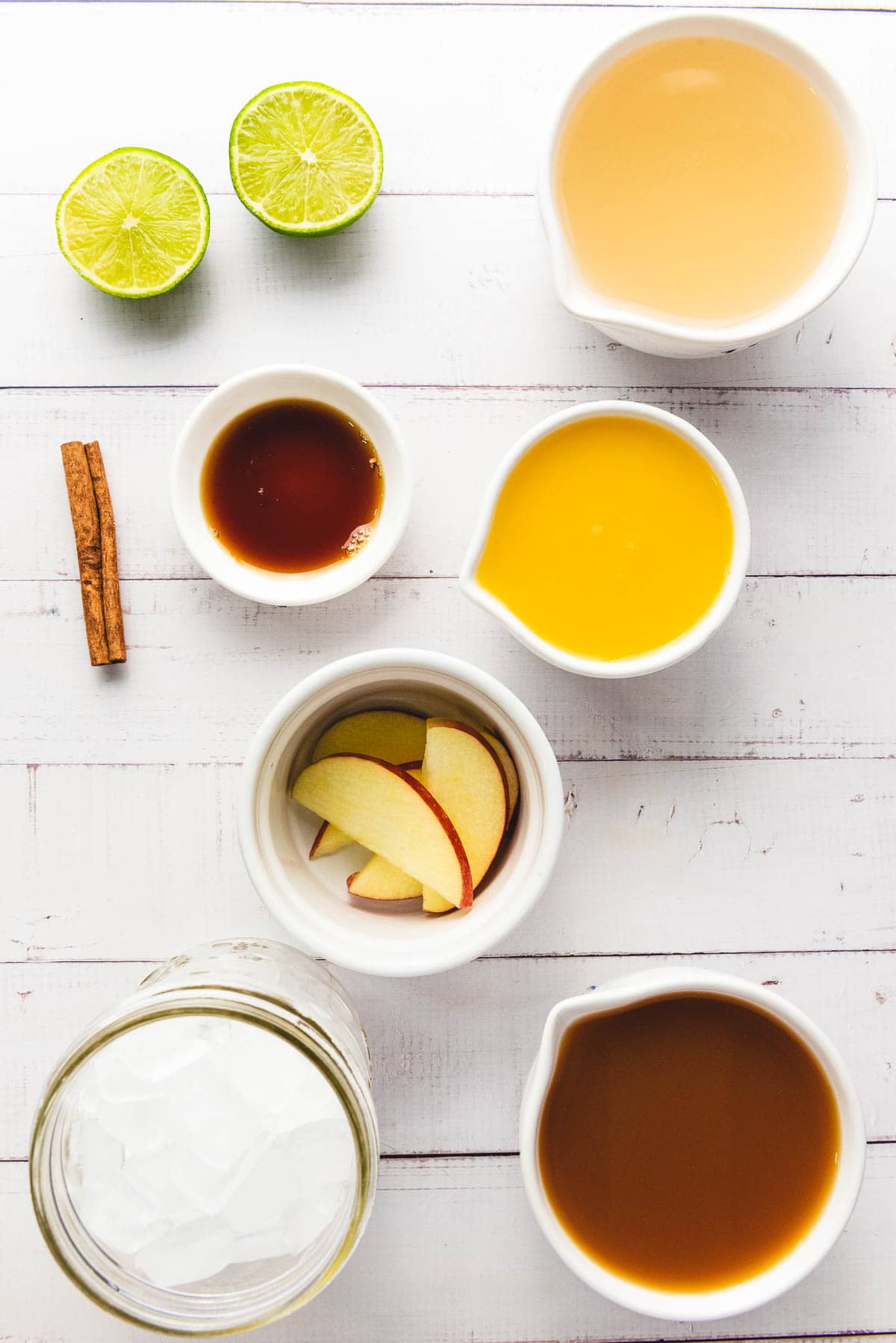 Top view of sliced lime, cinnamon sticks, apple slices, and cups of juice, tea, and apple cider on a white table.