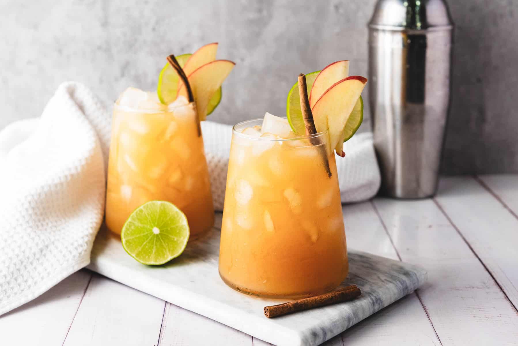 Two iced apple cocktails, garnished with apple slices and cinnamon, sit beside a lime and cocktail shaker on a marble board&mdash;perfect for serving as an elegant apple cider mocktail.