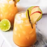 Two glasses of iced orange mocktail garnished with apple slices, lime, and cinnamon sticks on a marble board.