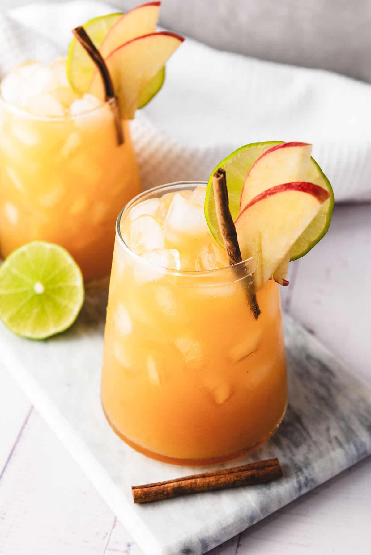 Two glasses of iced orange mocktail garnished with apple slices, lime, and cinnamon sticks on a marble board.