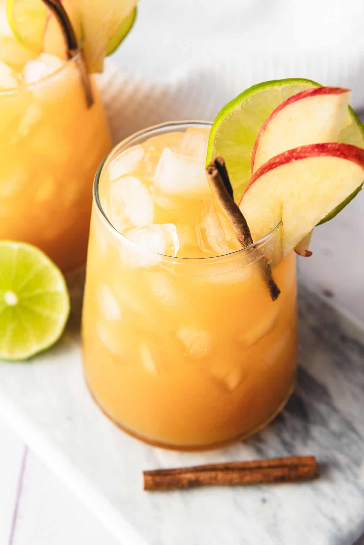 A refreshing mocktail featuring a glass of iced orange drink, topped with apple slices, a lime wheel, and a cinnamon stick&mdash;perfect to enjoy with a splash of ginger beer for added zing.