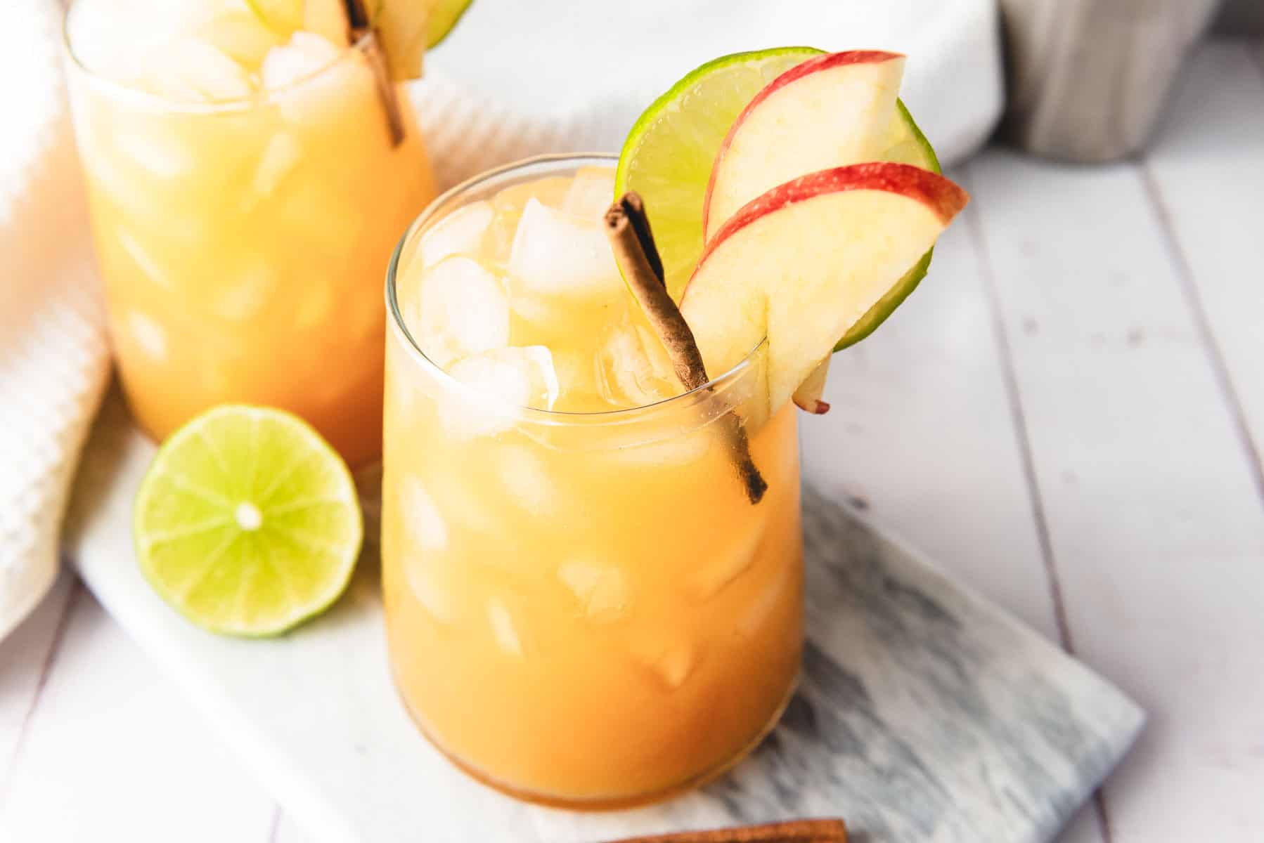 A glass of iced orange mocktail garnished with apple slices, lime, and a cinnamon stick on a marble board.