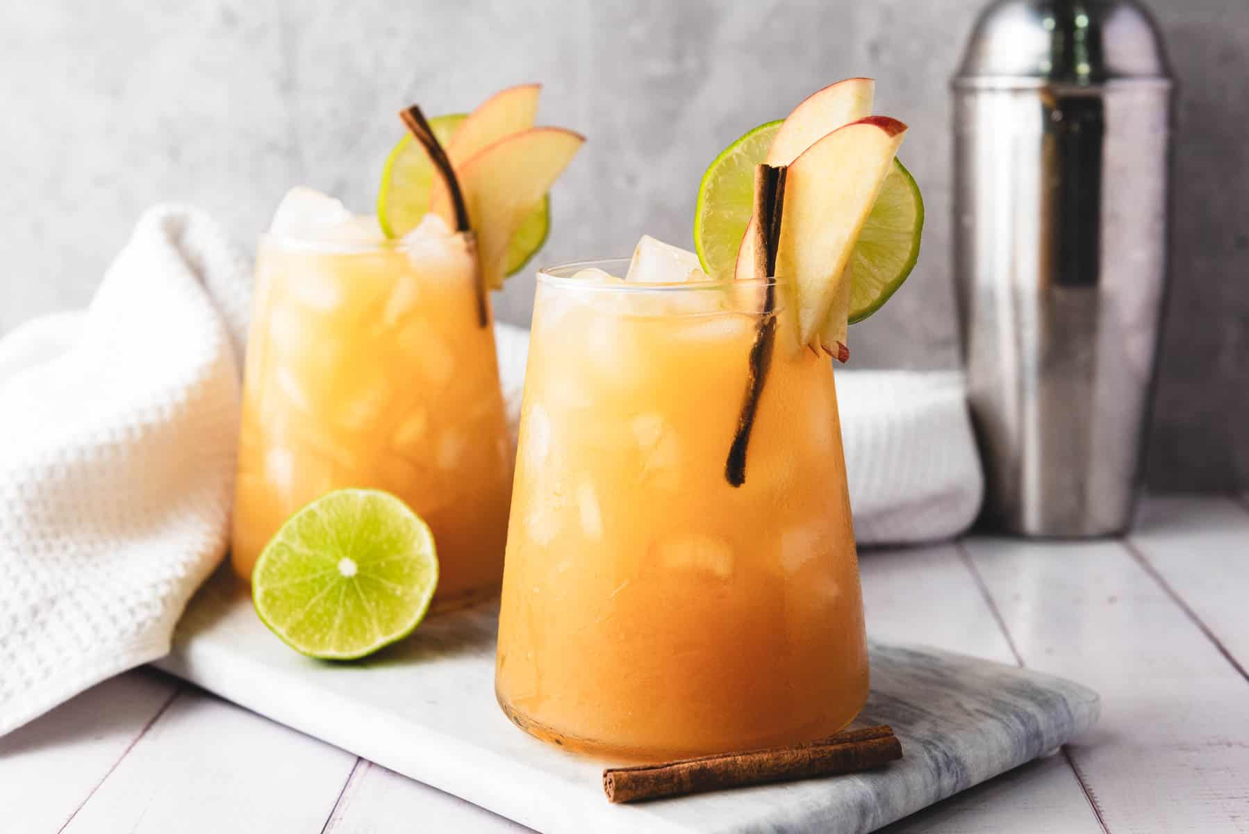 Two iced apple cider mocktails, garnished with lime slices, apple slices, and cinnamon sticks, are served on a marble board&mdash;perfectly refreshing with a splash of ginger beer.