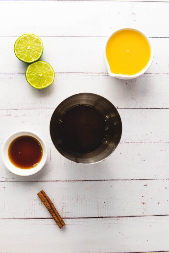 Overhead view of lime halves, orange juice, dark liquid in a shaker, vanilla extract, a cinnamon stick, and Ginger Beer on a white table&mdash;perfect for crafting a refreshing mocktail.