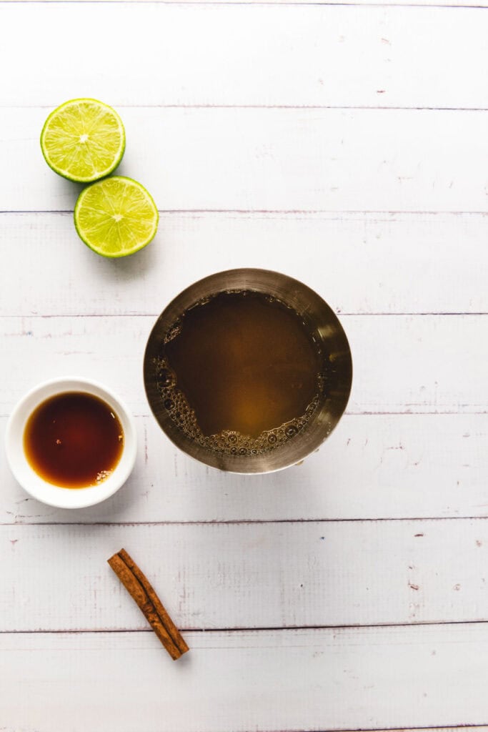 A metal cup of brown liquid&mdash;perfect for a mocktail&mdash;lime halves, a cinnamon stick, and a small bowl of syrup rest on a white table.