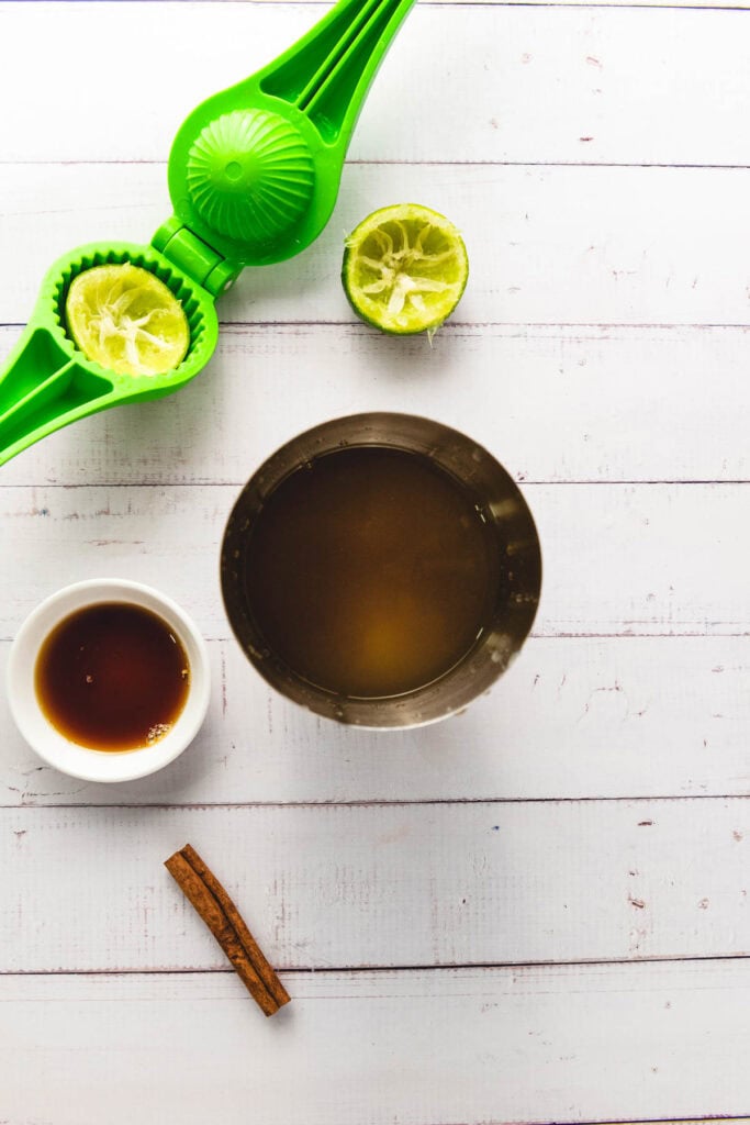 Metal cup with liquid, a cut lime in a green juicer, cinnamon stick, and small bowl of dark syrup on white table&mdash;perfect essentials for mixing an Apple Cider mocktail or adding a splash of ginger beer.