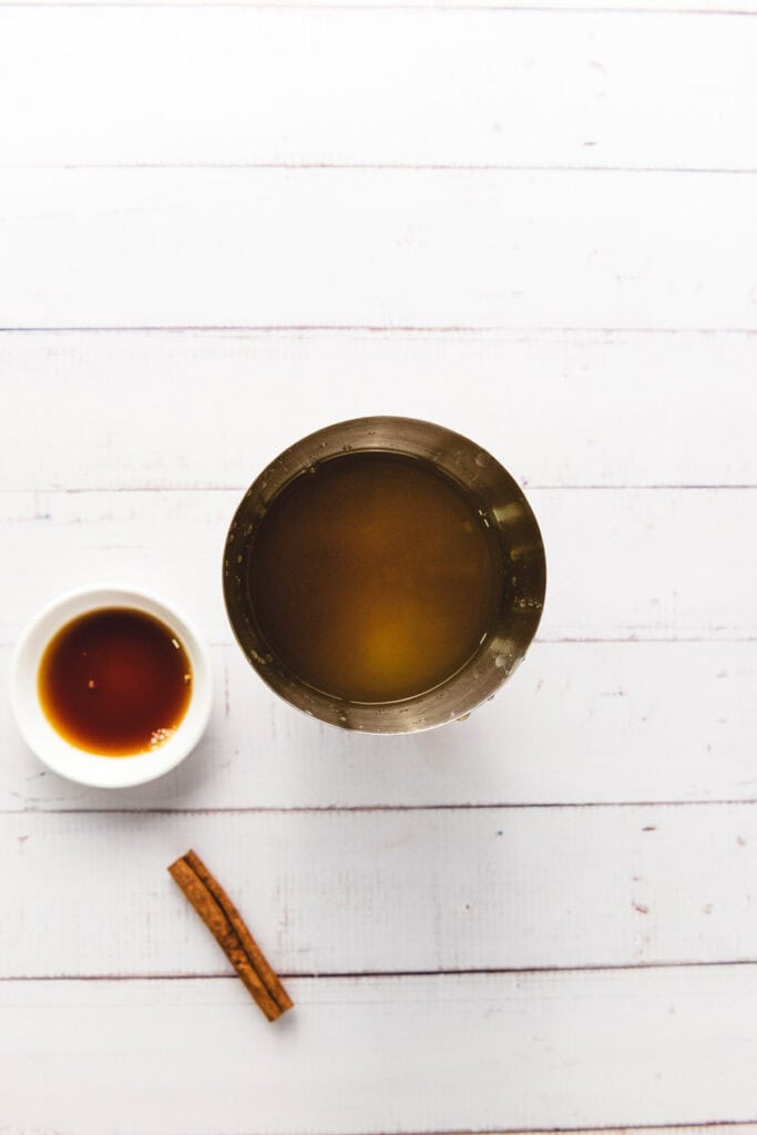 Overhead view of a metal cup filled with ginger beer, a small bowl of syrup, and a cinnamon stick on a white wooden surface.