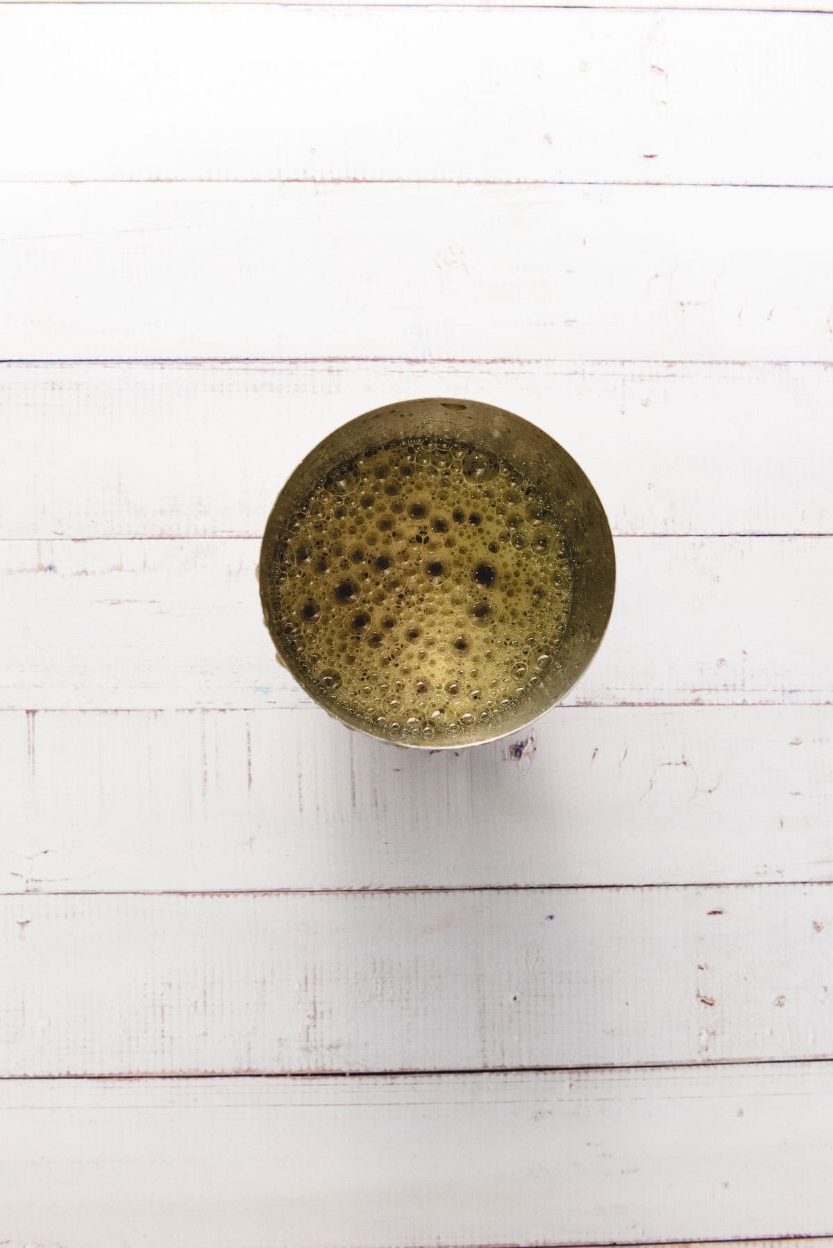 A round cup filled with a frothy dark apple cider on a white wooden surface, viewed from above.