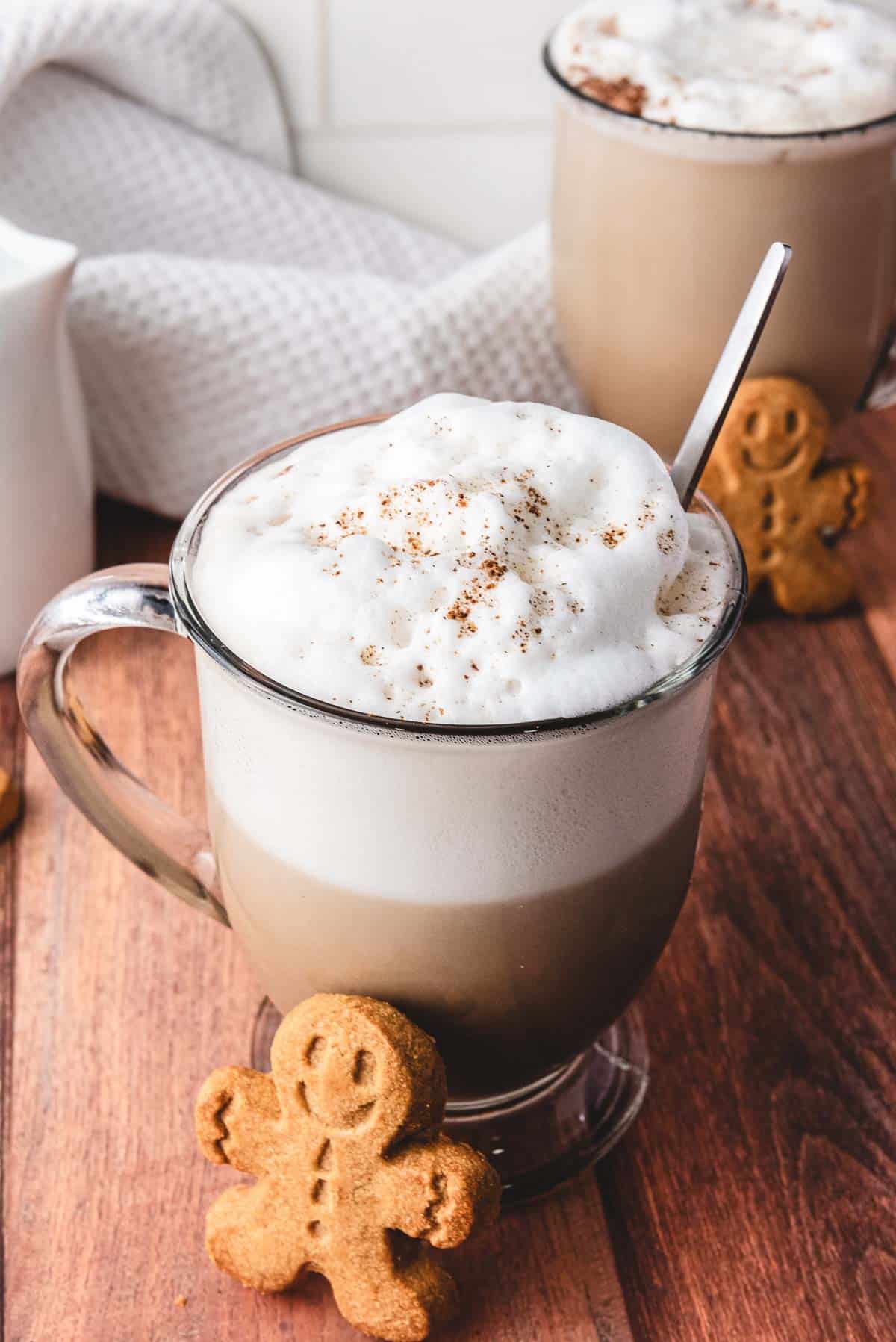 A cozy glass mug of frothy Gingerbread Latte with a gingerbread cookie on the side, set on a wooden table.