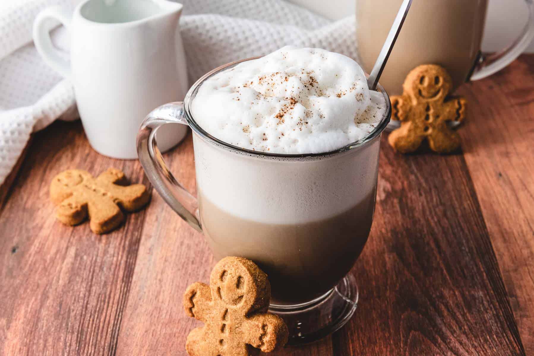 A cozy latte with frothy milk in a glass mug, surrounded by gingerbread cookies on a wooden table&mdash;a perfect Gingerbread Latte to enjoy with your favorite recipe.