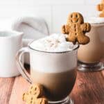 A glass mug of cozy latte with foam, topped with a gingerbread cookie, sits on a wooden table.