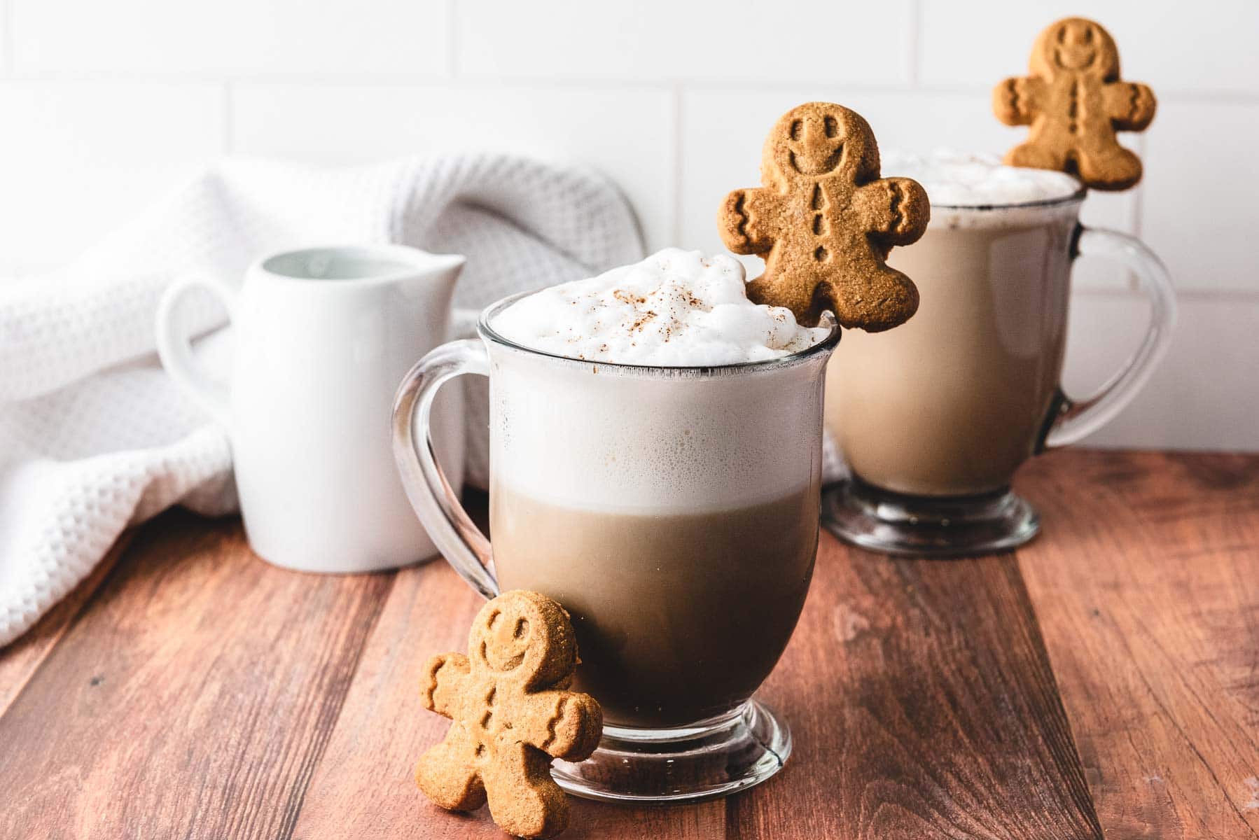 Two mugs of frothy Gingerbread Latte topped with cookies sit on a wooden table, creating a cozy scene with a white pitcher nearby.