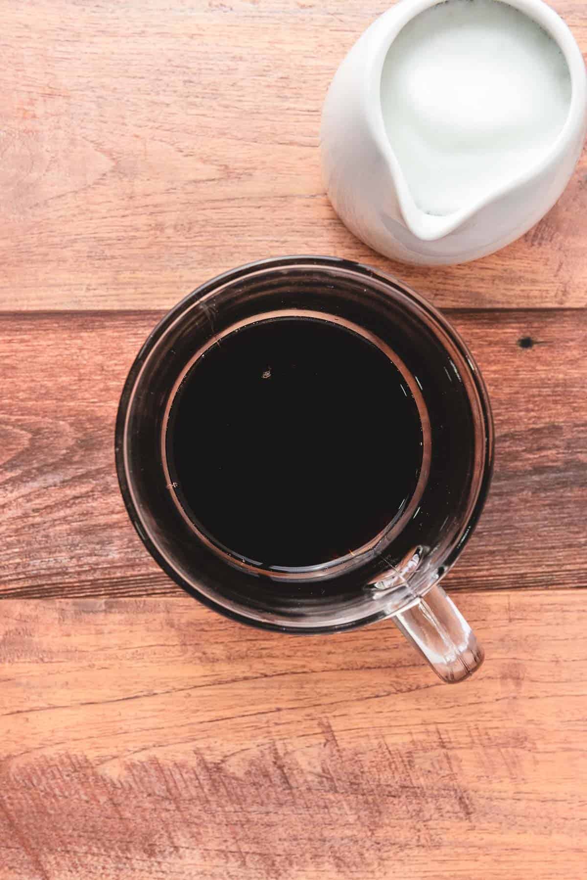 A cozy cup of black coffee and a small pitcher of cream sit on a wooden table, viewed from above&mdash;inviting inspiration for your next latte recipe.