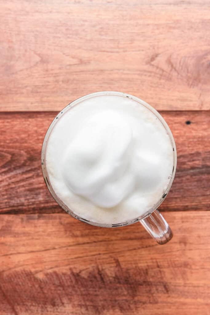 A glass mug of frothed milk on a wooden surface, viewed from above&mdash;perfect for crafting a cozy Gingerbread Latte recipe at home.